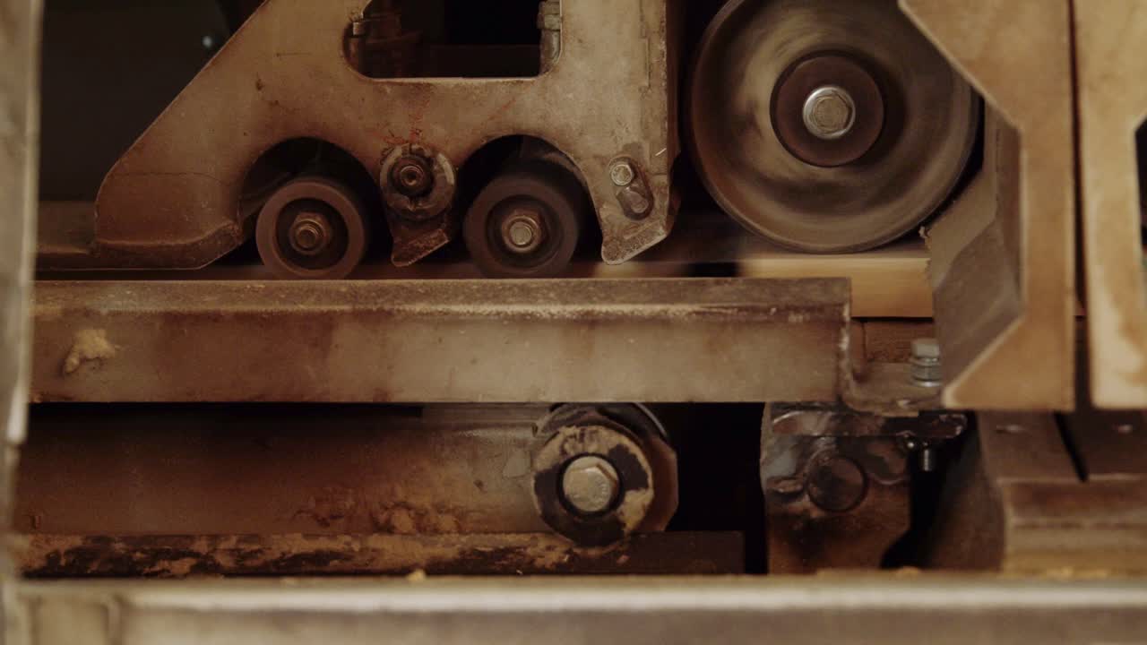 Macro detail of an industrial machine in a sawmill. Spinning rollers and wheels process a wooden plank, showcasing the power and precision of modern woodworking and manufacturing