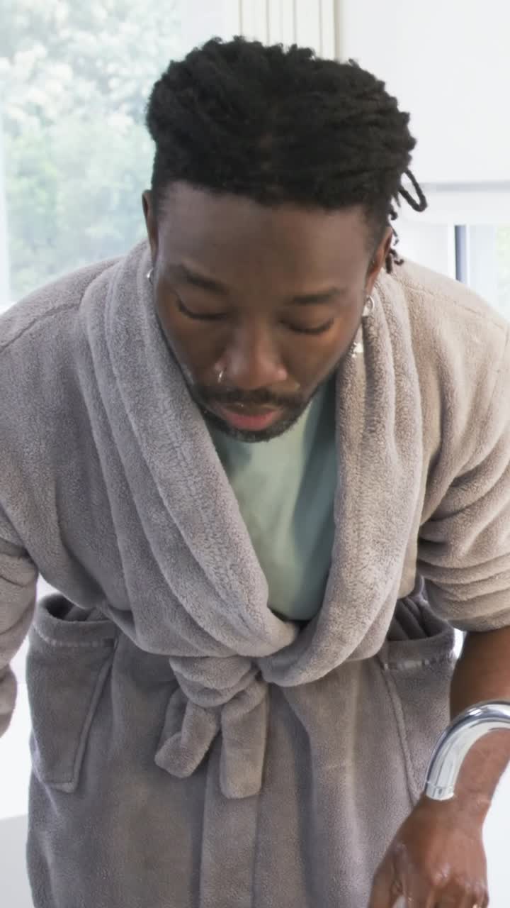 Vertical video: Leaning man in gray robe washing face at sink, rinsing and drying with towel