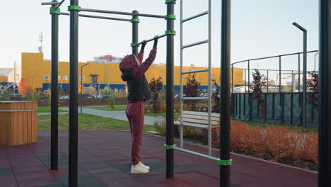 Side view female coach placing hands on pull up bar outdoors in urban park on rubber floor beside bench wearing burgundy hoodie vest before exercise demonstrating strength balance control