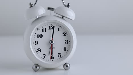 pequeño despertador blanco, números negros, fija la hora para las 18.00 horas, colocado en una mesa blanca.