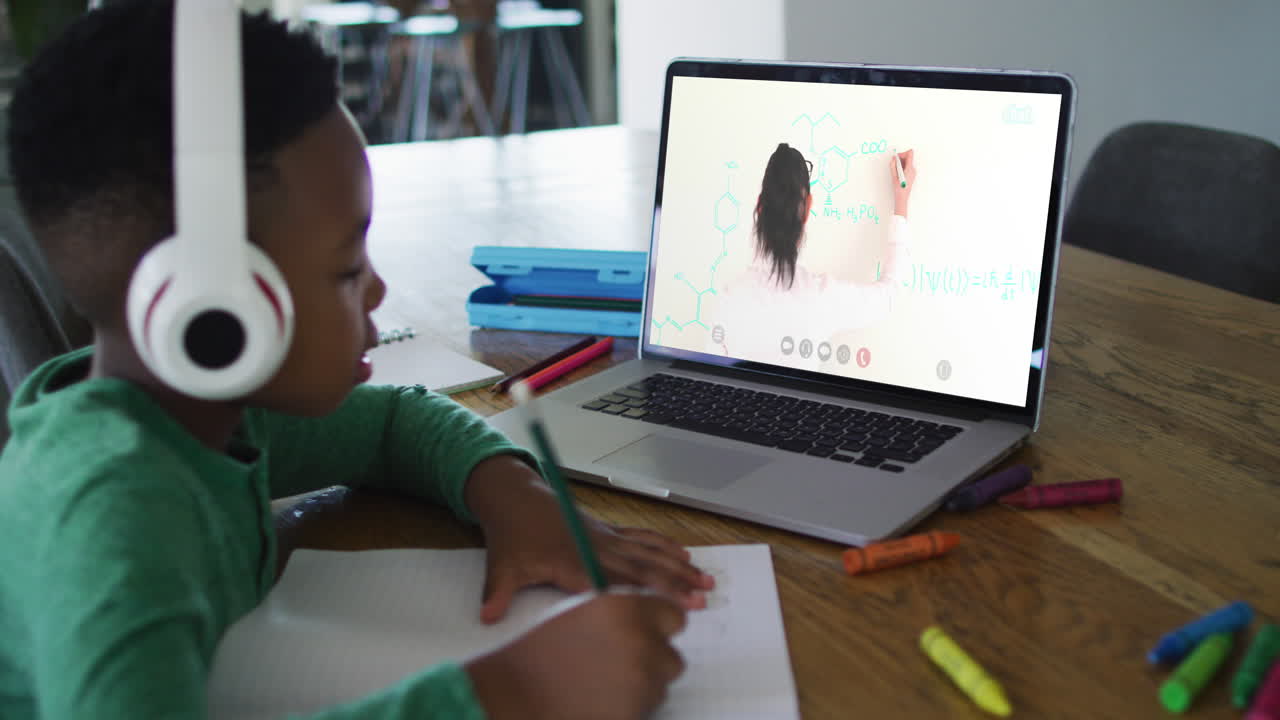 niño afroamericano haciendo la tarea mientras tiene una videollamada con una maestra en una computadora portátil en casa
