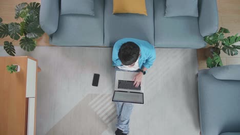 top view of a man thinking about something then raising index finger while typing on a laptop on the floor with smartphone in front of a sofa at home