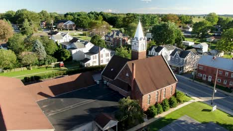 christian religion in america, united states small town with old brick church and steeple along street with neighborhood houses during summer, aerial orbit shot