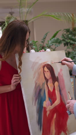 mujer mira el trabajo del artista en el caballete. hermosa mujer en vestido rojo sorprendida por la pintura maestra de la belleza en la tienda de arte. modelo comprueba el dibujo de retrato
