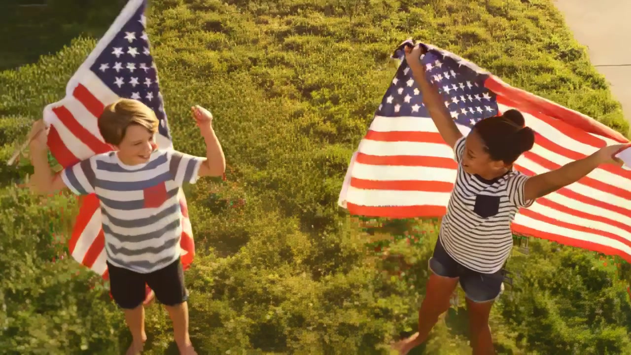 Waving two kids responding to overhead camera recording in front yard, posing with large US flags
