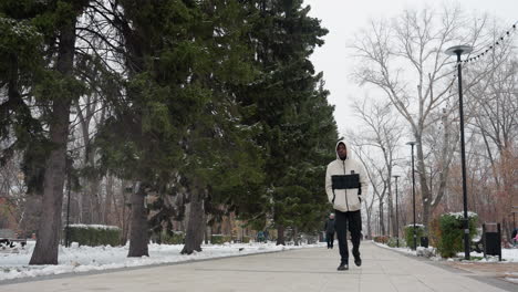 black man in white and black jacket with hands in pockets strolling on snowy park pathway chewing gum, bare trees, evergreen trees, benches, streetlights, and pedestrians in background