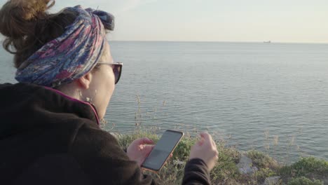 Eine-Frau,-Die-Zigarette-Raucht-Und-Mit-Dem-Telefon-Auf-Dem-Hügel-Mit-Meerblick-Spielt