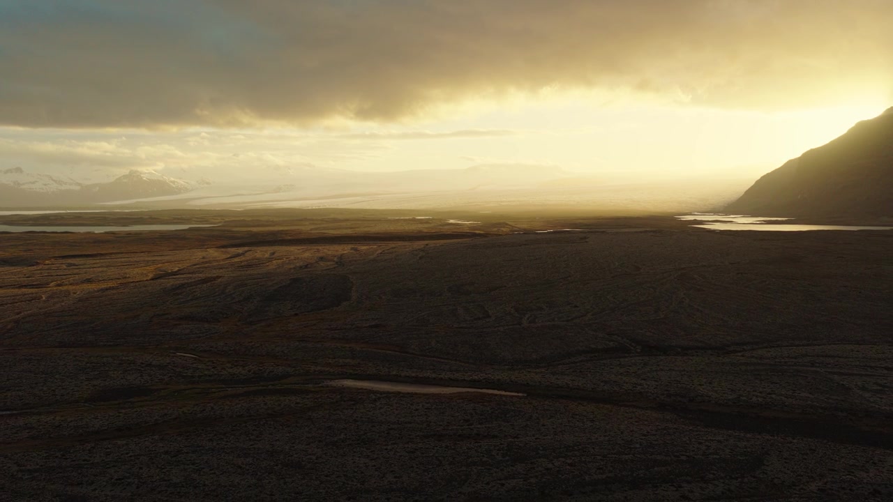 Burfell volcanic terrain and river channels stretch across a dark highland plain in South Iceland, illuminated by low evening sunlight beneath heavy clouds, drone pullback.