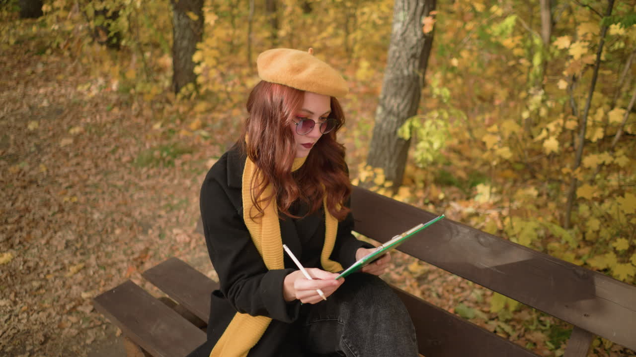 творческий ум вдумчиво рисует на холсте, сидит с наклоненной головой влево на деревянной скамейке в парке, погруженный в художественное выражение, одет в желтый берет и шарф