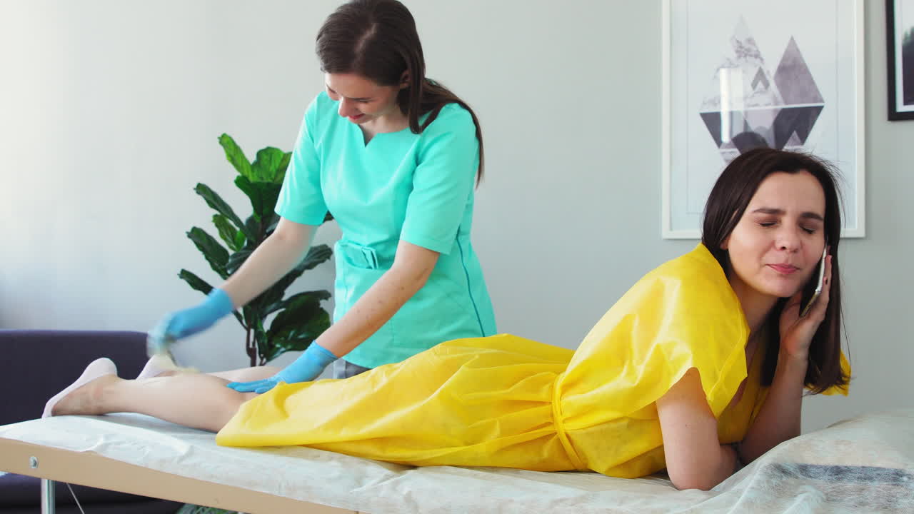 la chica habla por teléfono y sonríe durante la depilación de sus piernas. el maestro de shugaring elimina el pelo de las piernas
