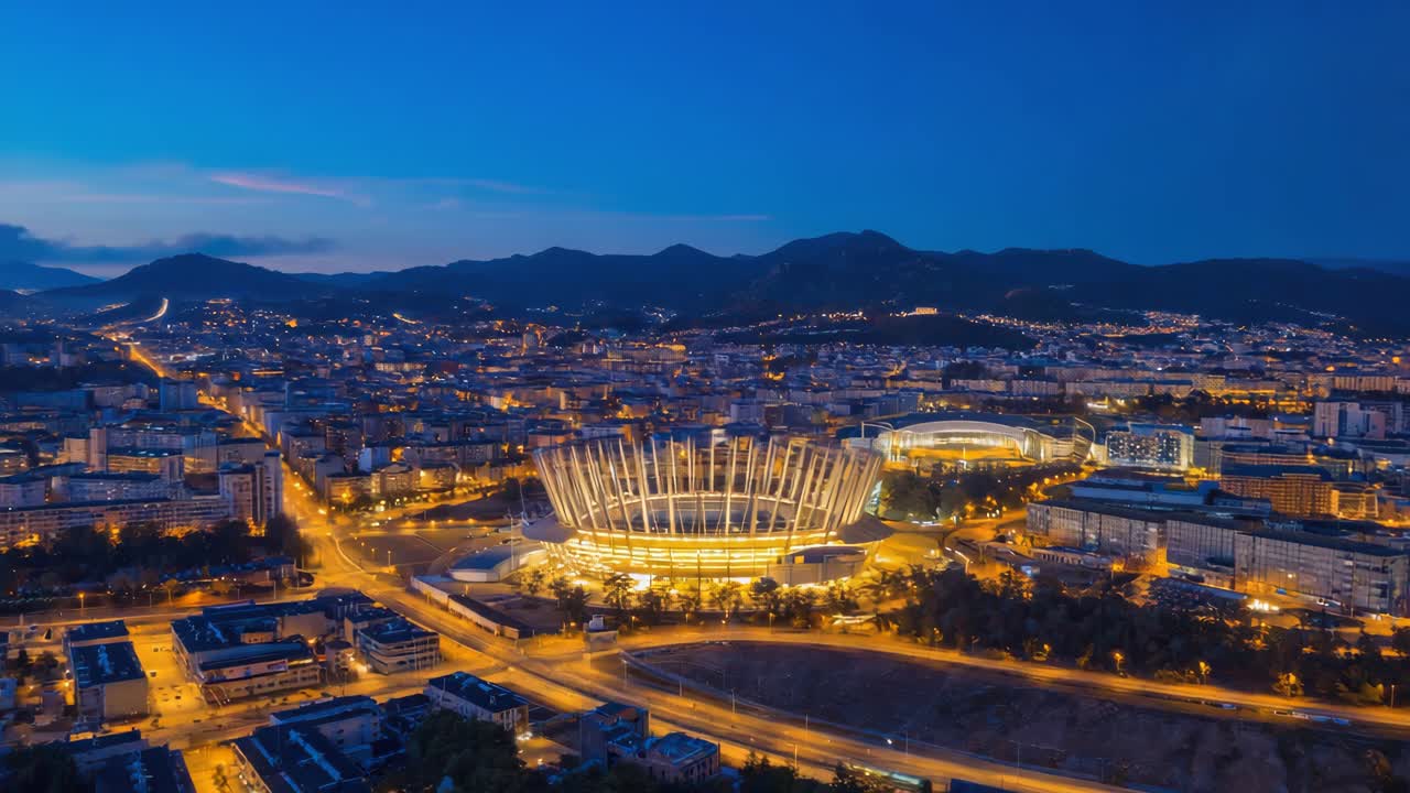 Aerial night view of a modern illuminated stadium in a vibrant city with mountains in the background