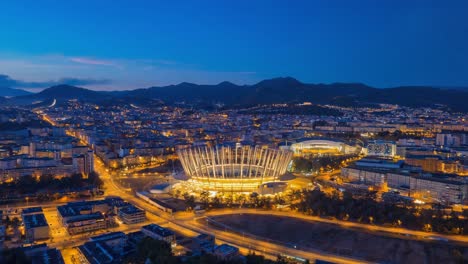 aerial night view of a modern illuminated stadium in a vibrant city with mountains in the background