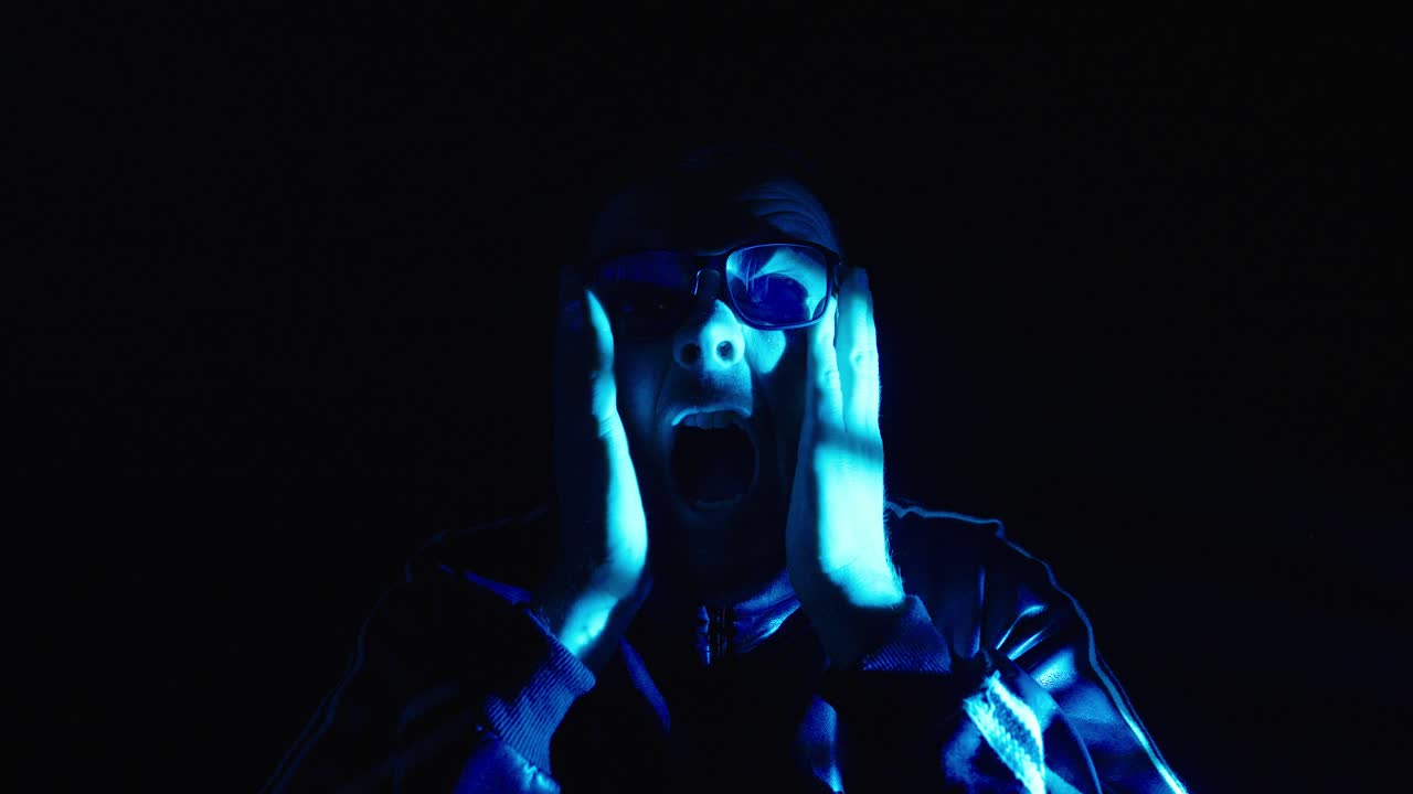 Primo piano cinematografico di un uomo che urla direttamente nella telecamera sotto una luce blu, creando un'atmosfera inquietante in stile horror con intensa paura e panico.