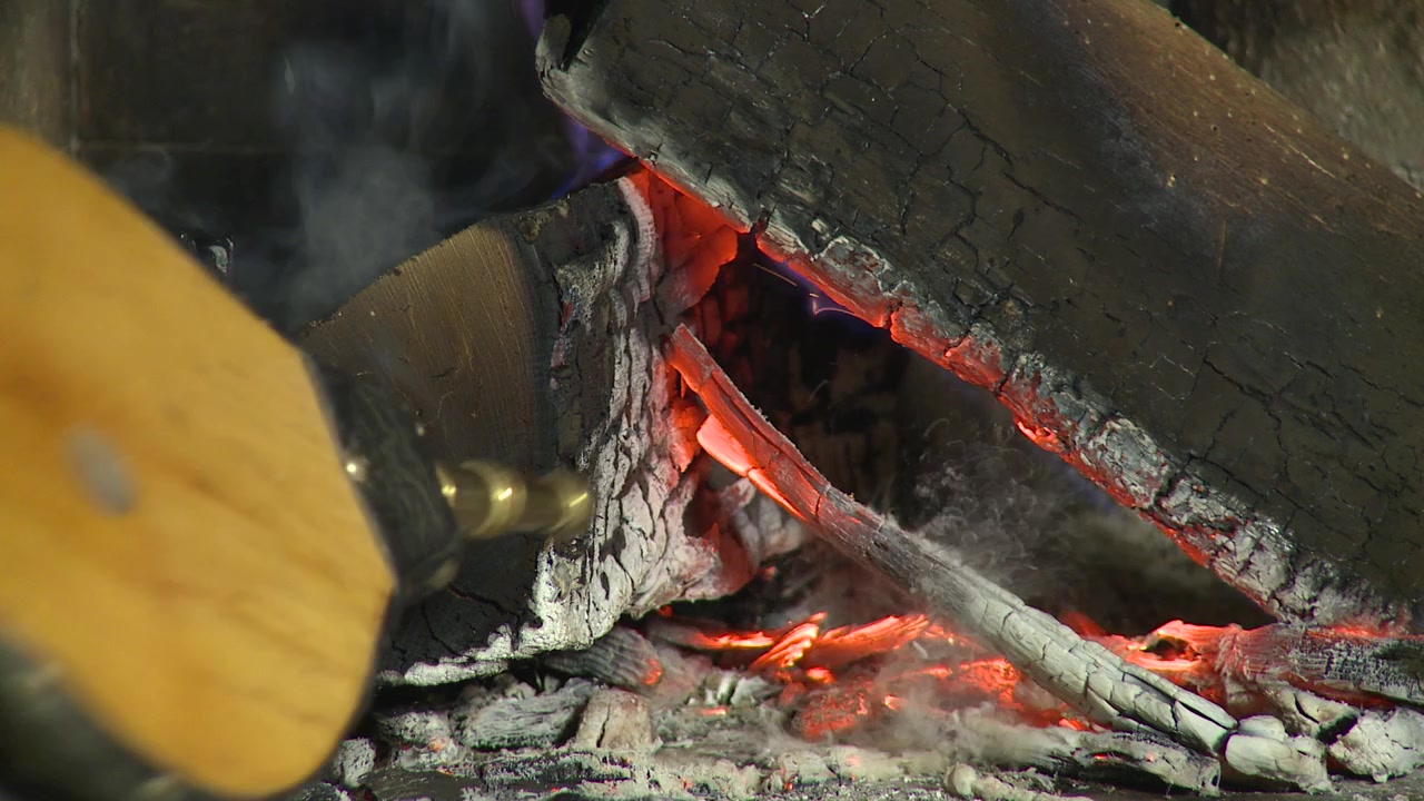 Close-up de um fole de lareira soprando ar sobre brasas incandescentes e lenha em chamas, reavivando um fogo de lenha quente em casa