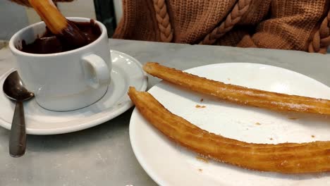 Female-hand-grabbing-some-churros-from-a-plate-and-dipping-it-in-a-mug-of-thick-dark-hot-chocolate