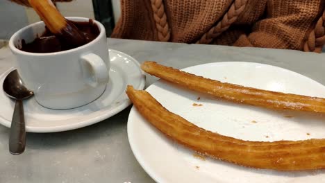 female hand grabbing some churros from a plate and dipping it in a mug of thick dark hot chocolate