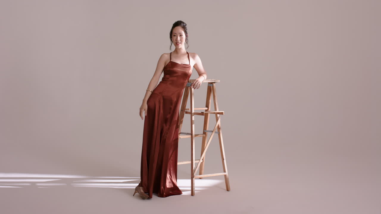 Chinese female model entering studio, leaning on ladder, pivoting back, brushing cheek in light