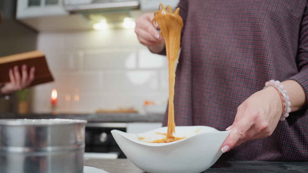 Close-up of woman in plaid dress lifting thick caramel-colored batter with whisk as it slowly drips back into white bowl, kitchen background with soft lighting, baking tools, and blurred cookbook