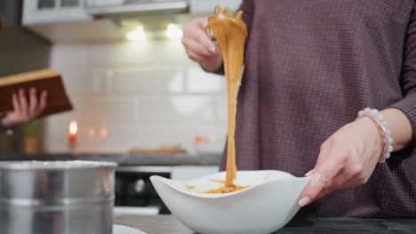 primer plano de una mujer con vestido a cuadros levantando una masa gruesa de color caramelo con un batidor mientras cae lentamente de nuevo en un cuenco blanco, fondo de la cocina con iluminación suave, herramientas de horneado y libro de cocina borroso