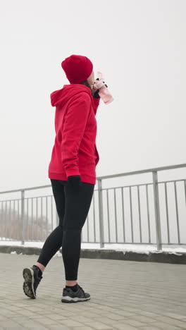 lady in red hoodie and beanie sips water from pink bottle outdoors in serene winter park setting with snow-covered ground, benches, trees, iron railing, and misty atmospheric background
