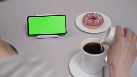 woman sitting at table coffee donut using smartphone with chroma key green screen, scrolling through social network media online shop internet. smartphone in horizontal mode with green screen mock-up.
