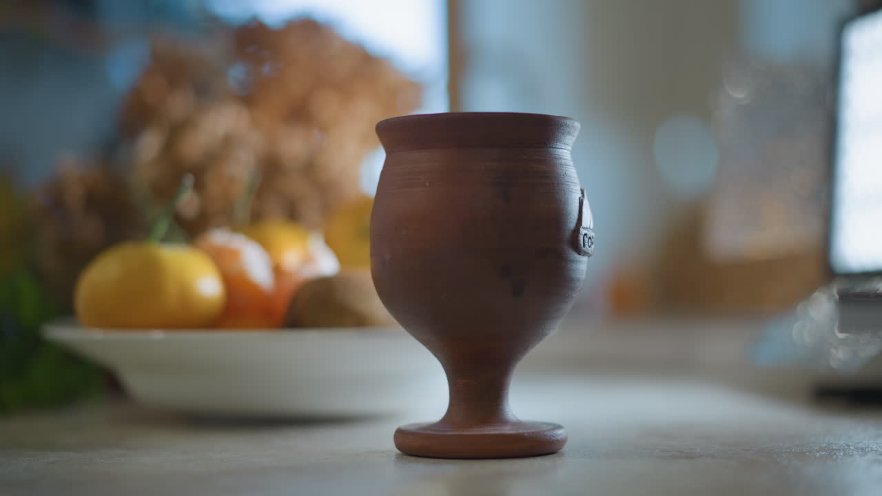 Copa rústica de arcilla en la mesa de la cocina. Luz solar suave, cuenco de cítricos y flores secas, quietud en primer plano con un sutil bokeh, ambiente cálido de cocina casera, superficie texturizada y poca profundidad de campo.