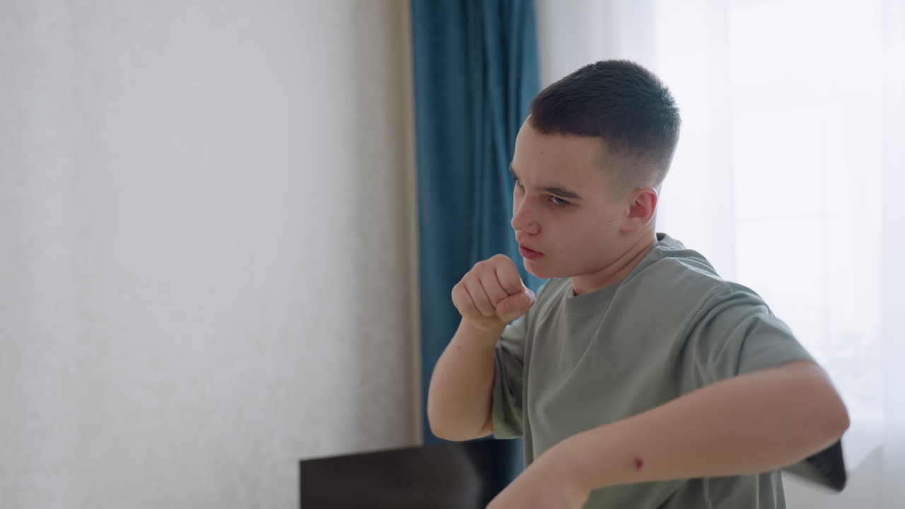 Focused teen boy rehearses boxing technique indoors with raised fists and concentrated gaze, wearing casual clothes in bright room with curtain backdrop