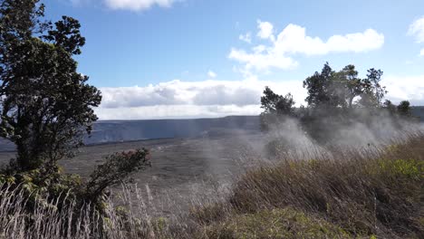 Steam-exits-the-ground-where-vegetation-grows-at-a-safe-distance-from-the-Kilauea-volcano-crater-in-the-background-on-a-typical-sunny-day-with-blue-skies-and-clouds