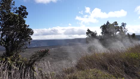 el vapor sale del suelo donde crece la vegetación a una distancia segura del cráter del volcán kilauea en el fondo en un típico día soleado con cielos azules y nubes