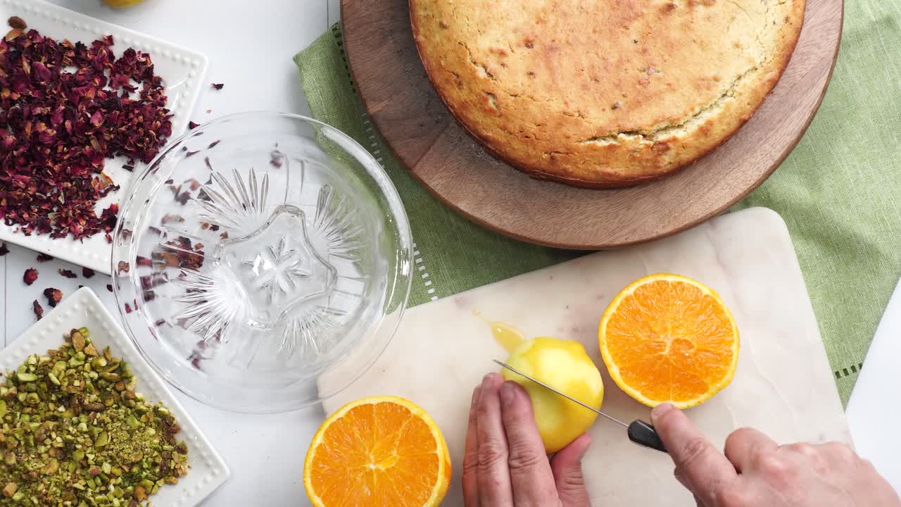 Baking a dessert called Persian Love Cake made with rose petals and pistachios. Cake with white glaze frosting. Cutting an orange on a cutting board. Beautiful cake for tea time or celebration