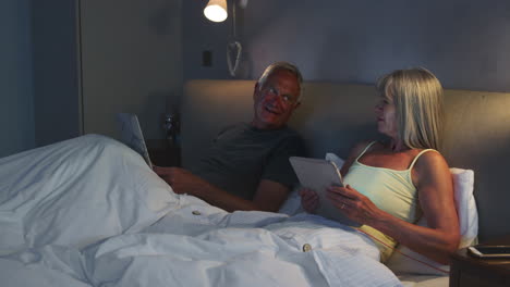 senior couple wearing pajamas lying in bed using digital devices