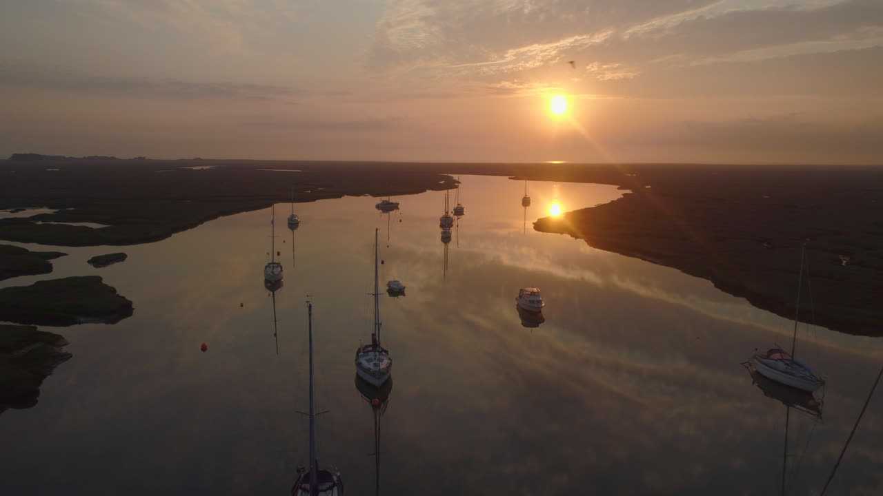 만조 때 개울에 있는 범선의 낮은 영화 같은 드론 샷, 새들이 날아가는 모습, 멋진 오렌지색 일출을 배경으로 반사된 채 노스 노퍽 영국 동부 해안