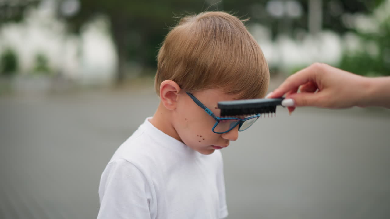 un niño con gafas se sienta en silencio mientras la mano de una mujer le peina el cabello, el niño parece cansado, con un fondo borroso de árboles y otros objetos poco claros