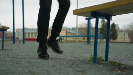 Close-up of a person's legs jogging slowly, wearing black jeans and black sneakers, near a bench with green grass underneath, a painted yellow house is visible in the distance