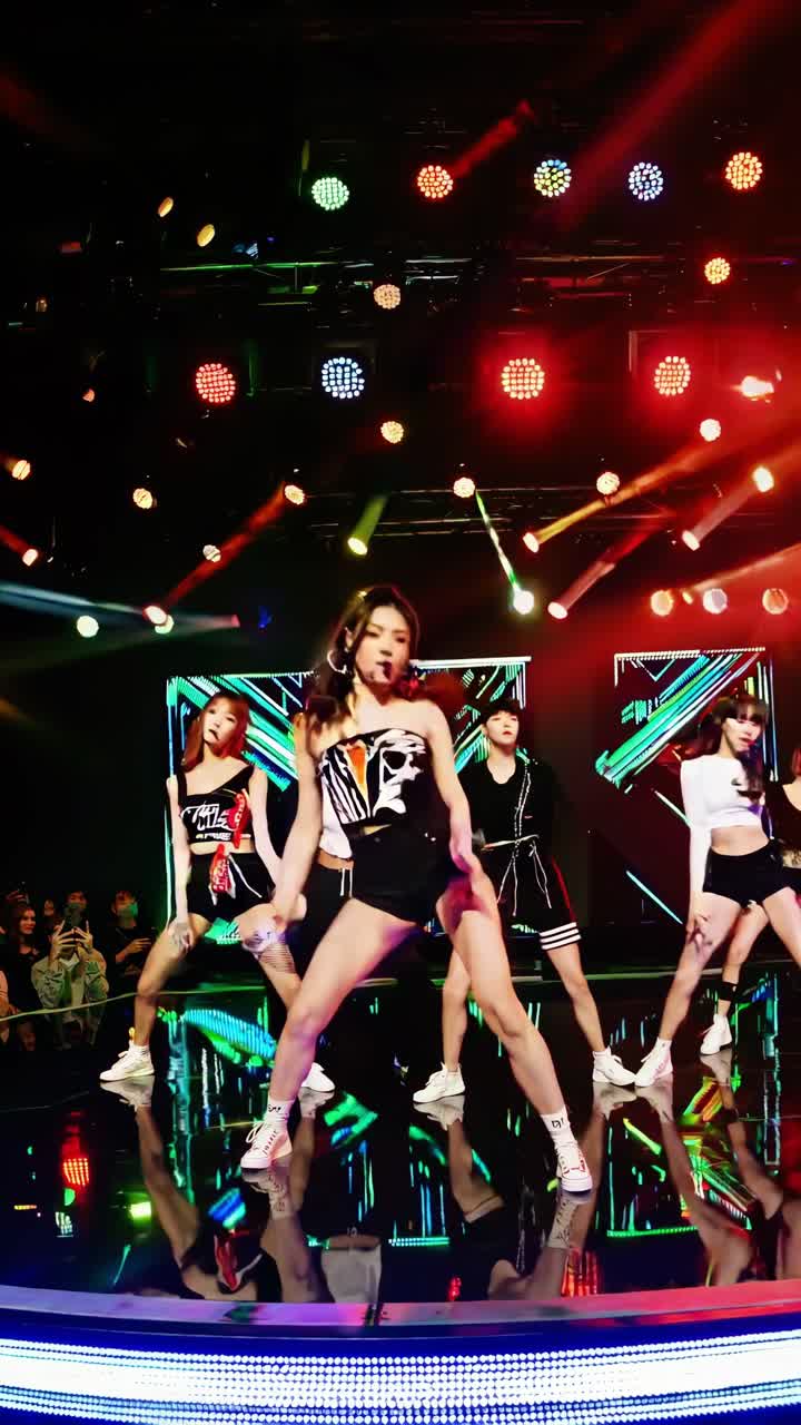 Energetic kpop group performing a dynamic dance routine on a brightly lit stage, showcasing synchronized movements and vibrant costumes under colorful spotlights and in front of a large led screen