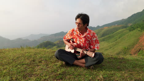 close up shot of an asian man hiker sitting on grass on top on a mountain and playing ukulele at sunrise