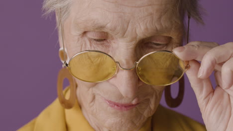 Nahaufnahme-Einer-älteren-Frau-Mit-Kurzen-Haaren,-Die-Ohrringe-Trägt,-Die-Mit-Den-Fingern-Auf-Einer-Sonnenbrille-Posiert-Und-Auf-Violettem-Hintergrund-In-Die-Kamera-Blickt