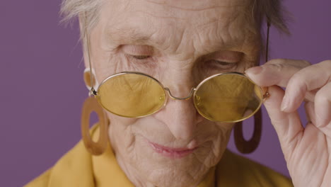 close up view of senior woman with short hair wearing earrings posing with fingers on sunglasses and looking at camera on purple background