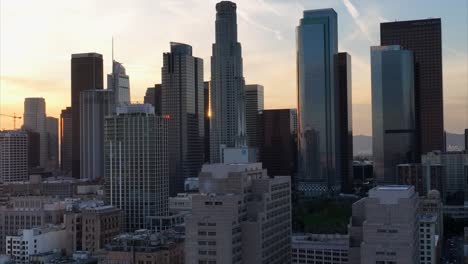 Skyline-Der-Innenstadt-Von-Los-Angeles-–-Aufsteigende-Luftaufnahme-Mit-Der-Untergehenden-Sonne,-Die-Zwischen-Den-Gebäuden-Leuchtet