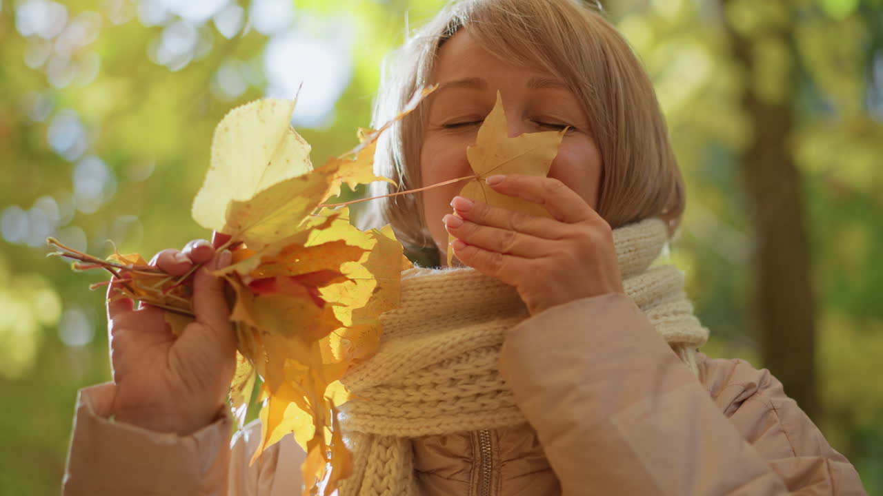 Lady smiling gently while admiring golden autumn leaves held in hands with sunlight creating soft glowing effect in vibrant forest setting filled with warm seasonal colors and blurred trees