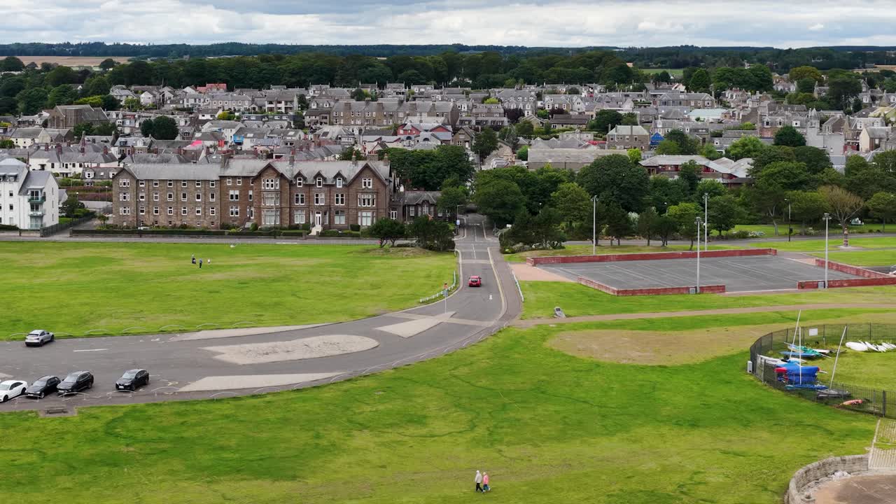 Drone glides from sandy beach over seawall to grassy park and town under cloudy daylight