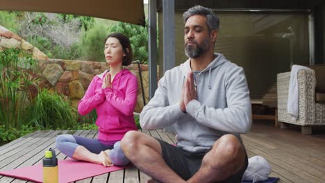 Happy-diverse-couple-doing-yoga,-sitting-and-meditating-at-terrace