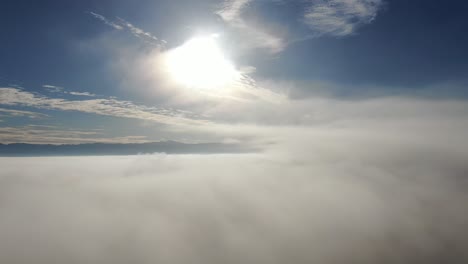 aerial flight by drone over a foggy landscape on a beautiful blue sky day passing through the mists with mottled clouds and sun above