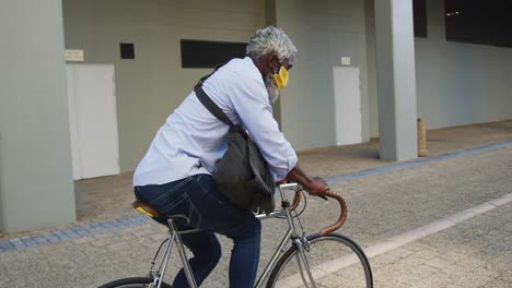 African-american-senior-man-wearing-face-mask-riding-bicycle-in-corporate-park