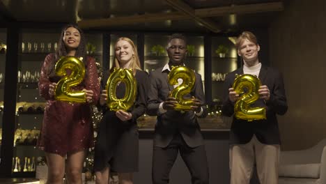 Group of friends wearing elegant clothes holding balloons with the numbers 2022 while dancing in new year&#039;s party