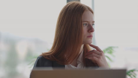 A-young-red-haired-business-woman-looks-thoughtfully-at-the-screen-and-brainstorms.-Watch-and-think-about-problems-looking-out-the-window.-Thoughtful-business-woman