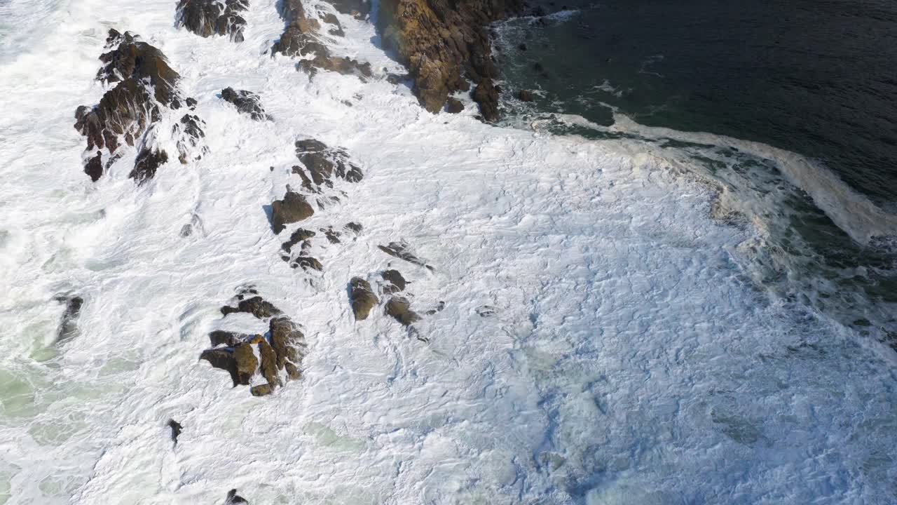 Drone footage captures powerful ocean waves crashing against rocky cliffs at Byron Bay, Australia, under clear daylight