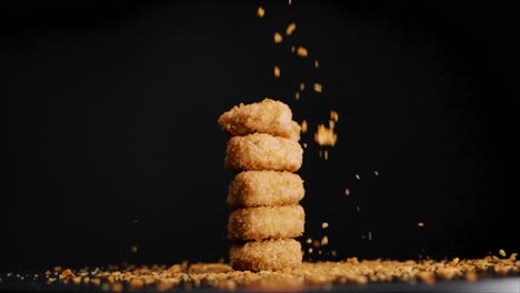 stack of vegan chicken nuggets with bread crumbs falling on them while they rotate, slow motion, black background, static shot