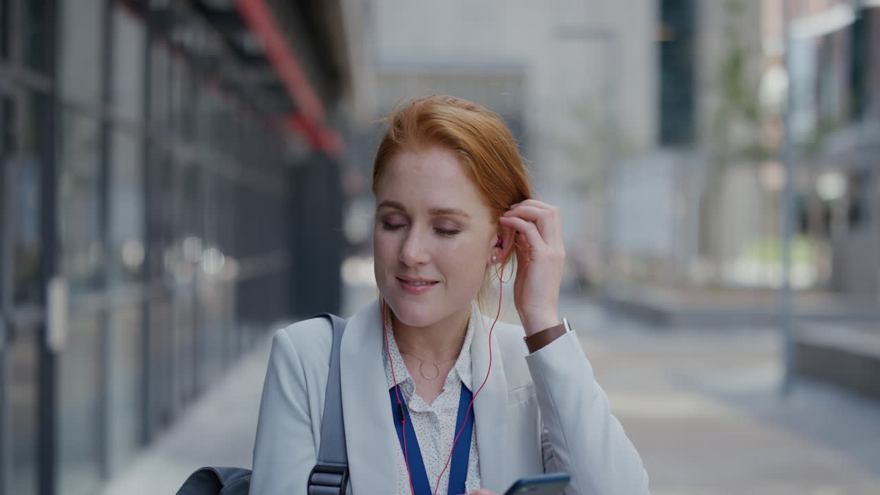 portrait happy young red head business woman using smartphone texting sending messages enjoying listening to music wearing earphones in relaxed urban city slow motion