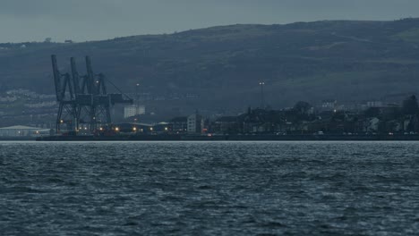puerto industrial con grúas al anochecer con aguas onduladas en primer plano